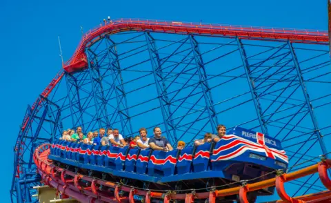 Pleasure Beach Resort A crowd of people on a rollercoaster, which features blue and red train carts. It is a summers day 