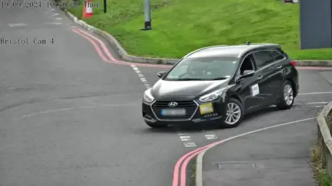 A CCTV camera shows Mr Shore's taxi at a junction near the airport. The car is black, and the road has red double lines running along it. 