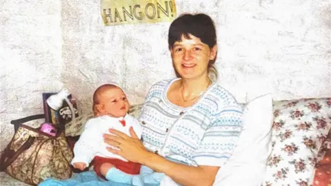 Family photo Linda smiles on a sofa while holding her baby daughter Cat. She has dark hair and wears a white and blue cardigan. Cat wears a white jumper, red shorts and long blue socks. 