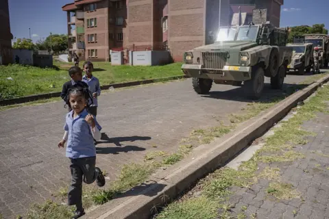 Anadolu via Getty Images Children in school uniform are seen running in front of an armoured vehicle as soldiers patrol in their neighbourhood