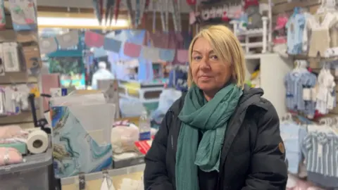 BBC/James Vincent Market trader at Halifax Market, Dawn Milner looks at the camera from her stall. She is wearing a padded coat and green scarf. She has blonde shoulder cropped hair. In the background are baby clothes hung up for sale.