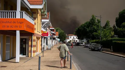 Pedestrians look at a cloud of black smoke covering the sky nearby of an area evacuated due to a wildfire in Lege Cap-Ferret.