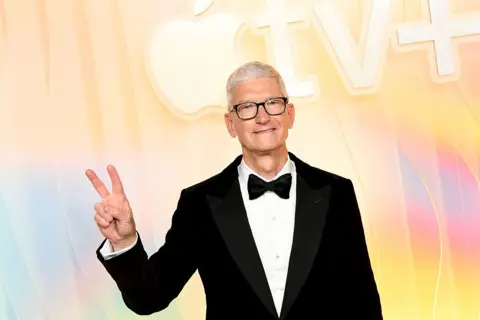 Getty Images Tim Cook wears a tuxedo and makes the "peace" sign as his stands in front of a yellow, blue and pink background reading Apple TV plus