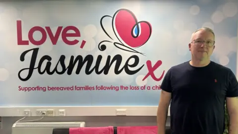 BBC Rob Lapsley, a middle-aged man with glasses, wears a t-shirt and stands in front of a large sign on a wall. The sign reads: 'Love, Jasmine. Supporting bereaved families following the loss of a child.'