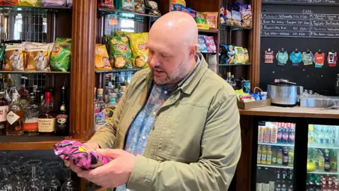 A man holding a pink packet of crisps behind a bar