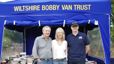 Wiltshire and Bath Air Ambulance Nick and Annette Mason standing with Wiltshire Bobby Van Trust Operator Scott Roughley under a blue gazebo with the words "Wiltshire Bobby Van Trust" on it and lots of books piled on tables underneath it.