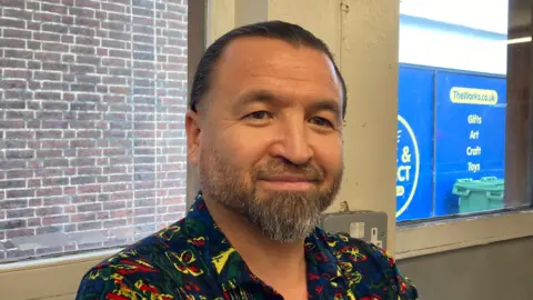 A man stood in front of a window. He has dark hair and a beard and is wearing a blue, red and yellow shirt. In the background is a red brick wall and a shop sign.