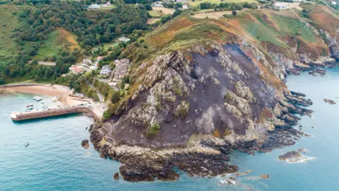 States of Jersey Fire and Rescue Service Aerial shot of a wildfire at Bonne Nuit. Smoke is billowing off the open area which has been damaged in the blaze. Water is surrounding the edge of the island. 