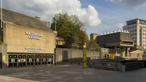 A large concrete-built building with the words "Southbank Centre" and "Queen Elizabeth Hall Purcell Room" on the side. There is a paved area in front with greenery to the side and a tower block in the distance.