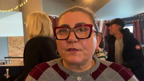 Woman with hair tied back, hoop earrings and black rimmed glasses wearing checked jumper 