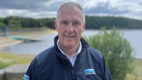 Paul Hudson/BBC Dave Kaye, who has short grey hair and blue eyes. He is wearing a blue Yorkshire Water zip-up jacket and he is standing in front of a reservoir.
