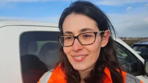 A picture a woman in glasses, wearing an orange hi-vis vest, with a wite truck behind her