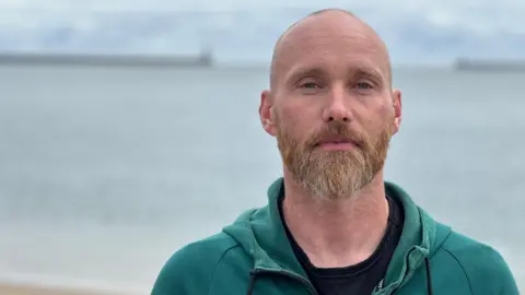 Olly Armstrong is standing with his back to the sea on a beach. He is mostly bald and has a thick beard and moustache. In the background, which is out of focus, is a pier with a lighthouse on it. He is wearing a teal coloured hoodie and black t-shirt.