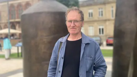 Chris is looking at the camera. He id wearing a navy blue over shirt and a black t-shirt underneath. He has thin framed glasses and short grey hair. 