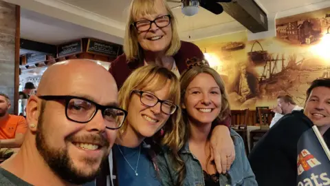 Family group picture of Linda Imrie smiling and standing behind her three adult children, who are sitting down in a restaurant.