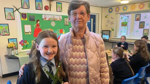 Carmel Hayes and her granddaughter Mya are standing together. Mya on the left and Carmel on the right. Mia is wearing school uniform and holding a green cross. Caramel is wearing a pink coat and jumper. They are in a classroom. There are more children in the background. 