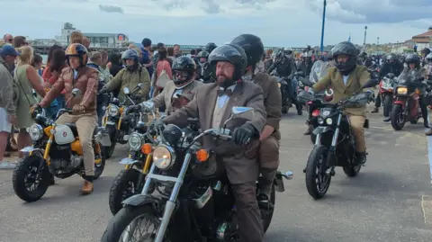 A group of motorbike riders are heading past the camera. They are all wearing similar brown suits. The closest rider to us has a brown bow tie, a beard, and is wearing glasses. 