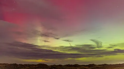 Christian Spooner Red, pink and green hues in the sky, dotted with cloud, above a landcape of grass covered sand dunes .
