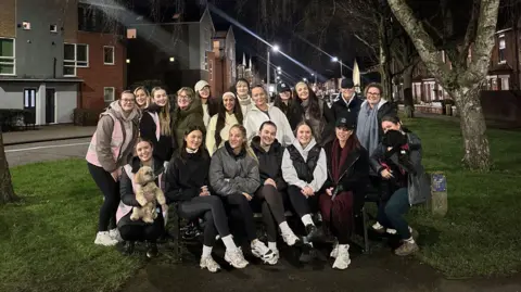 Girls That Hike A group of girls, some sitting on an outdoor bench and some standing up, smiling at the camera. It is night time. 
