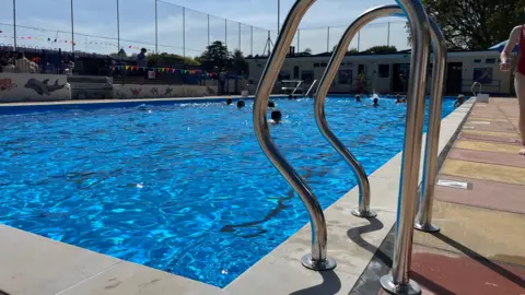 The refurbished pool under a blue sky. There is a set of metal steps in the foreground the about 15 swimmers in the water.