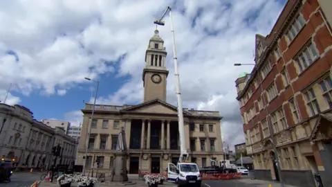 A tall, white crane on the back of a white van with a cage with engineering in it working above the city's Guildhall, an imposing sandstone building complete with columns and a clock tower.