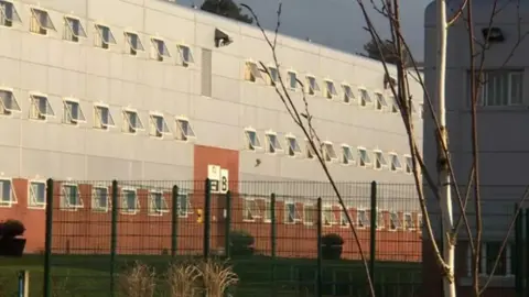 Parc prison from behind an green metal fence. the building is three stories and had brick on the main floor and silver metal cladding on the second and third floors. 