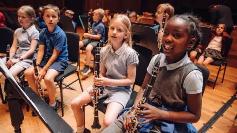 Children from Horfield CE Primary school at Earthsong day at Bristol Beacon, 2024. The children are in school uniform style outfits and the main children - four of them, are all holding clarinets. They have music stands in front of them and are all sitting as if ready to begin playing. 