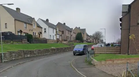 A road going up a hill with houses on either side. The houses on the left side are raised on a hill with grass in front of them and bordered by a stone wall. Cars are parked along the side of the road.