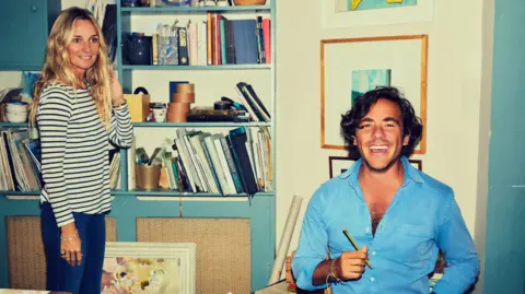 Chris Floyd Jack Savoretti laughing during a photoshoot at his home. He is sitting down and is holding a pencil. His wife Jemma Powell is standing near him, near a bookshelf, full of books and other items.