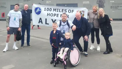 Ashley A group of people stand outside a grey building with a banner held between them which says "100 miles of hope for Alyssa". In front is a girl with blonde hair in a pink wheelchair with a boy standing to her right.