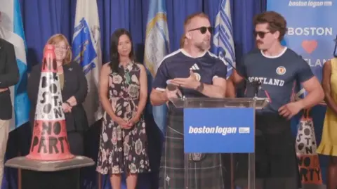 A traffic cone with the message "No Boston, no party" is seen on the left, while two men in kilts and sunglasses are seen behind a podium on the right.