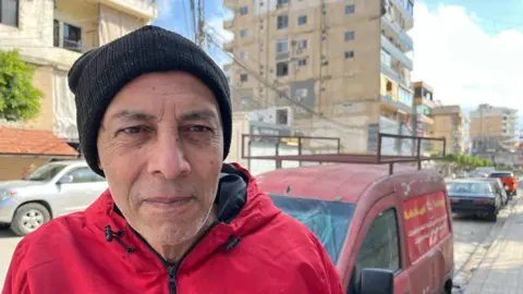 An older man looks into the camera, expression neutral, wearing a black hat and red jacket on the street.
