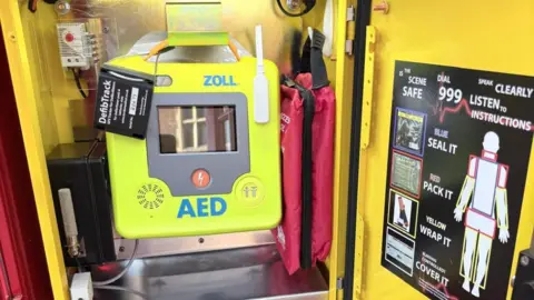 Matt Gaskin A bright yellow automated external defibrillator unit mounted inside the phone box alongside medical kits and a poster showing step-by-step emergency instructions.