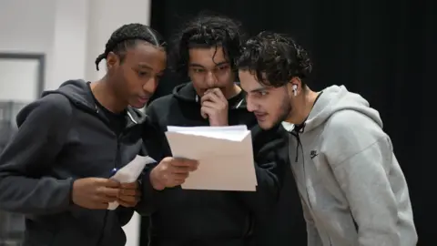 Three young men anxiously read the one in the middle's A-level results. He is holding his hand up to his mouth. 