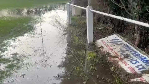 A heavily waterlogged area beside a football pitch with standing water submerging the grass. A white perimeter railing and a partially visible advertising board are lying on the ground.