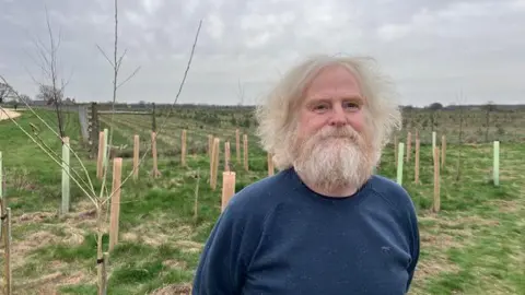 A man in a blue top is smiling at the camera with a field behind him that has been planted with small trees, some of the trees have protective orange and green tree tubes around them.