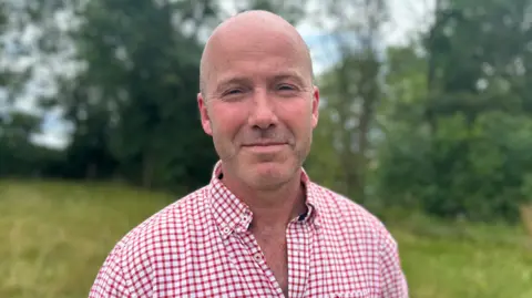 Charles Goadby is wearing a red-and-white check shirt. He is bald with short stubble on face.