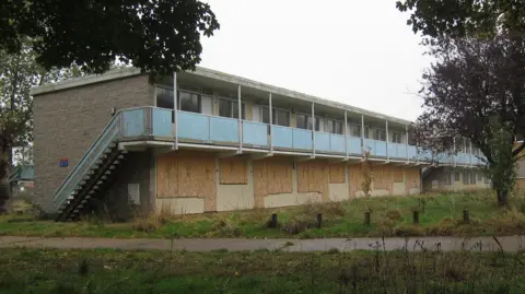 Geograph/Hugh Venables A two-storey building is derelict. The bottom floor's windows are boarded up. A blue painted staircase leads to the second floor. There is grass in the foreground.