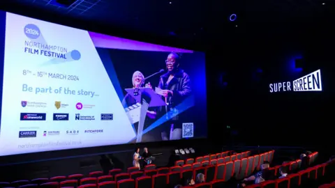 Northampton Film Festival A cinema-like room featuring a large screen highlighting the Northampton Film Festival and rows of red seats. Details of the event and 11 sponsors are listed on the screen. There are red seats in the auditorium. 