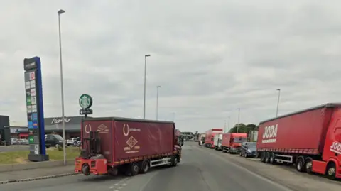 A red lorry turns onto the A49 with a line of traffic on the opposite carriageway led by another larger red lorry