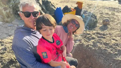 Image of a white haired man, wearing sunglassses, digging a hole on a beach with a young boy sitting on his lap holding a beach spade and a woman with a sunhat on and sunglasses sitting in the background.