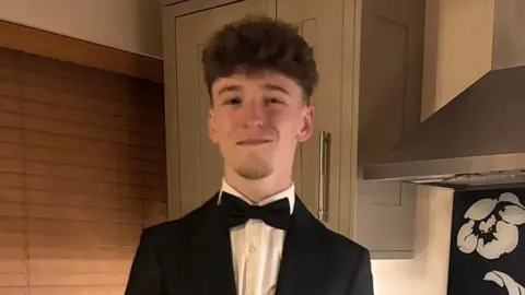 Family photo A teenage boy stood in a formal suit, including a black jacket and black bow tie, stood in a kitchen, smiling for the camera.