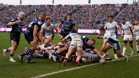 PA Media Bath's Sam Underhill leans over the line to score a try with one hand for Bath against Gloucester at the Rec. Multiple Bath players are behind watching on while Gloucester players in their all-white change kit try and stop the try