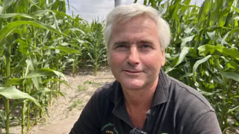 Jeremy Stokes smiles as he sits at a table with tall maize plants growing around him. He has white hair and is wearing a black polo-neck top.