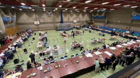 Ben Schofield/BBC A sports hall has been set up for election counting. Tables are set up in a horseshoe shape with some other tables in the middle. Lots of people are sitting at the table or standing looking over. 