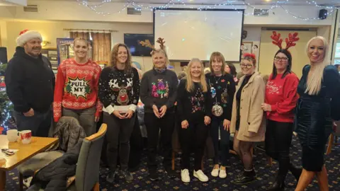 Romsey Royal British Legion Nine people wearing Christmas hats and jumpers stand in a row inside the Royal British Legion in Romsey. Mary Hillman, on the right, has long blonde hair and wears a dark dress.