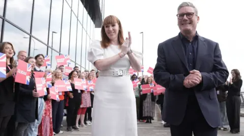 PA Media Sir Keir Starmer, wearing a dark suit, smiles next to Angela Rayner, dressed in white, as the two arrive in Liverpool ahead of Labour's Party conference. Against the backdrop of the two are Labour supporters who are smiling, applauding, cheering and holding up red Labor signs.