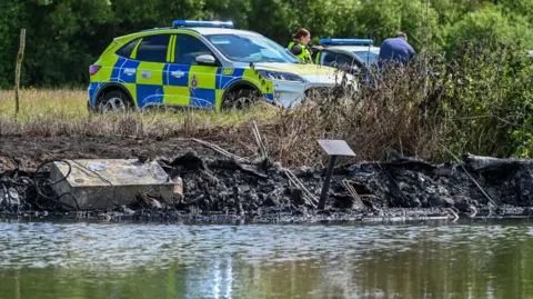 SWNS Two police cars parked by the river bank, with an officer stood by the car. In the foreground the charred wreckage of the boat can be seen in the water.