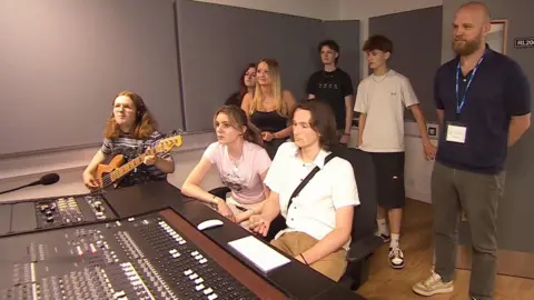 Will Champion stands to the right of the image, he is bald with a short beard and wears a navy blue polo top with grey jeans and beige and white Nike dunk trainers. He is standing in a recording studio with a group of students.