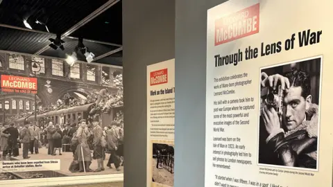 LEONARD MCCOMBE A board with a picture of McCombe holding up his camera with the words Through the Lens of War above it, with a view through to the exhibition on the right.